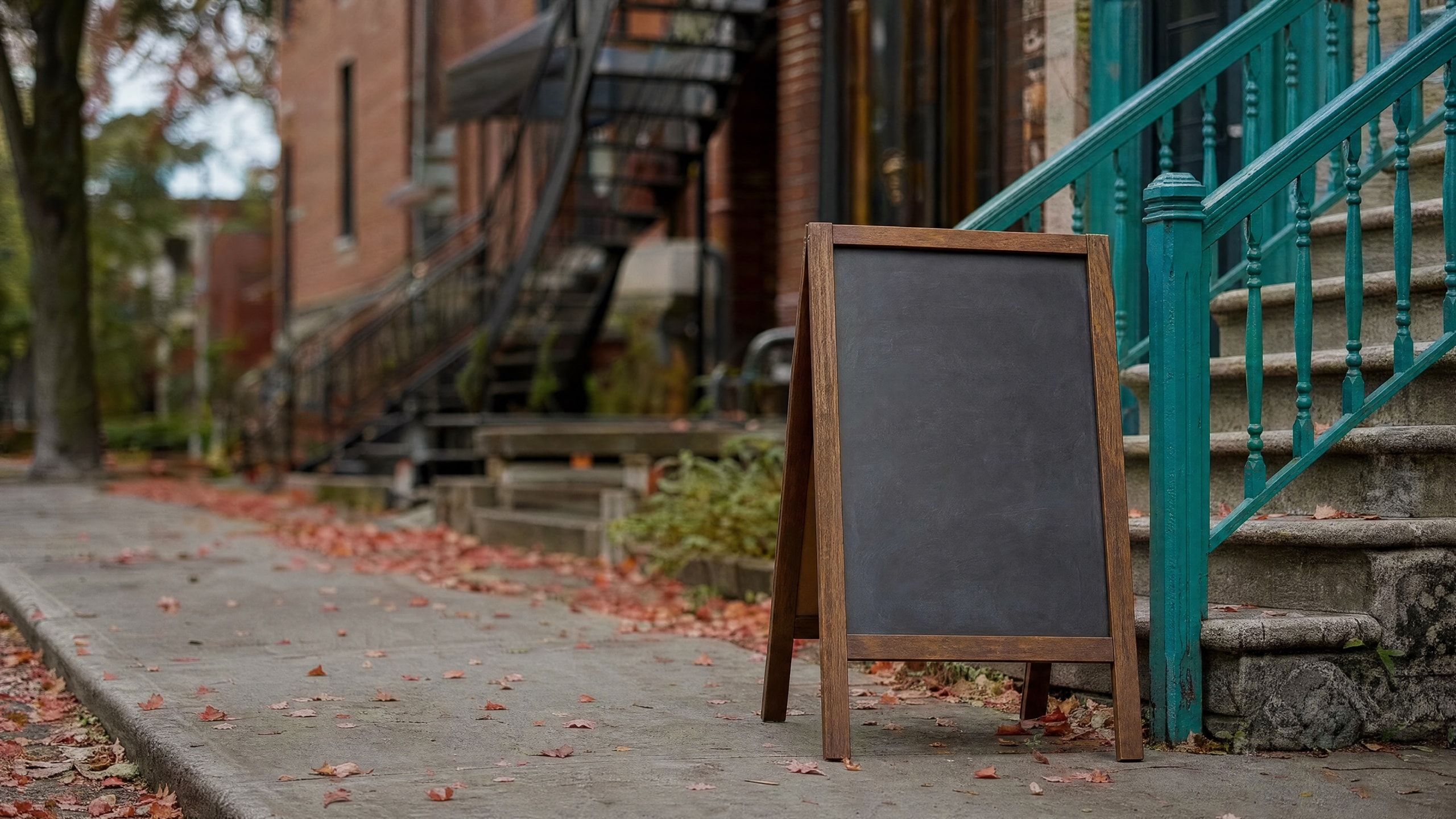 Une enseigne-chevalet à ardoise effacée, sans aucun mot, sur un trottoir de Montréal en automne, devant des triplex de brique et leurs escaliers de fer.